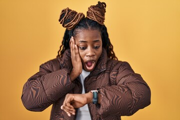 African woman with braided hair standing over yellow background looking at the watch time worried, afraid of getting late