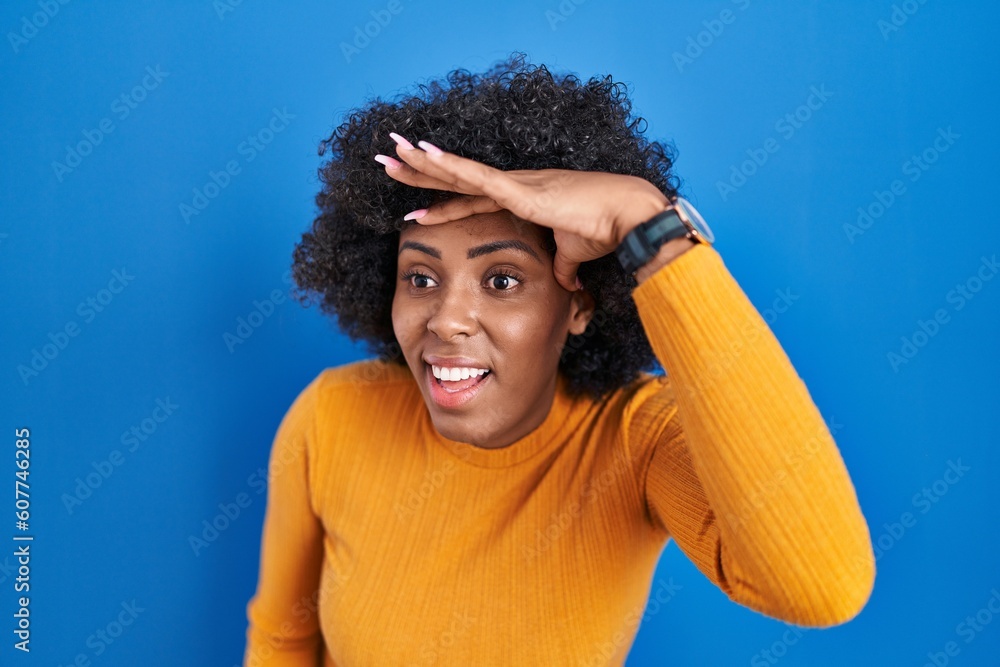 Poster Black woman with curly hair standing over blue background very happy and smiling looking far away with hand over head. searching concept.