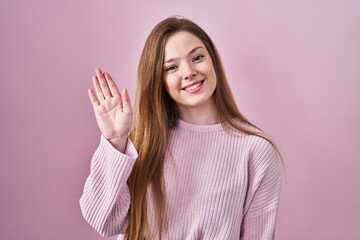 Young caucasian woman standing over pink background waiving saying hello happy and smiling, friendly welcome gesture