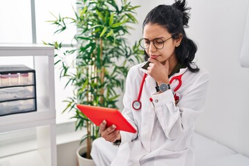 Young latin woman wearing doctor uniform using touchpad with doubt expression at clinic