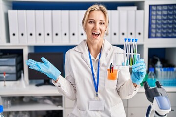 Young caucasian woman working at scientist laboratory holding test tubes celebrating achievement with happy smile and winner expression with raised hand