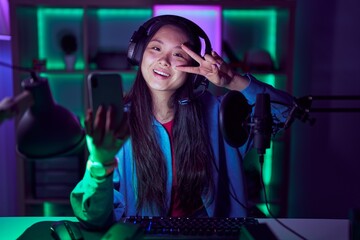 Obraz na płótnie Canvas Young asian woman playing video games with smartphone doing peace symbol with fingers over face, smiling cheerful showing victory