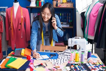 Fototapeta premium Young chinese woman tailor talking on smartphone writing on notebook at clothing factory