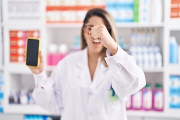 Blonde woman working at pharmacy drugstore showing smartphone screen annoyed and frustrated...