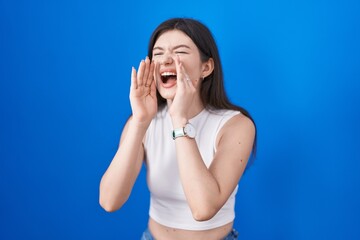 Fototapeta premium Young caucasian woman standing over blue background shouting angry out loud with hands over mouth