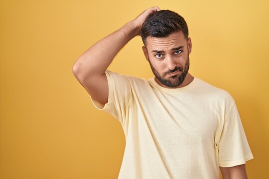 Handsome hispanic man standing over yellow background confuse and wondering about question. uncertain with doubt, thinking with hand on head. pensive concept.