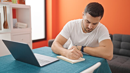 Young hispanic man using laptop taking notes at dinning room