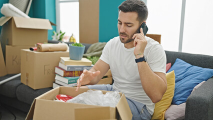 Young hispanic man unpacking cardboard box talking on smartphone at new home