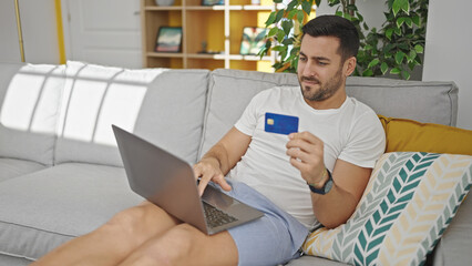 Young hispanic man shopping with laptop and credit card sitting on sofa at home