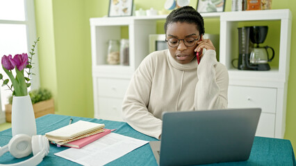 African american woman using laptop talking on smartphone at dinning room