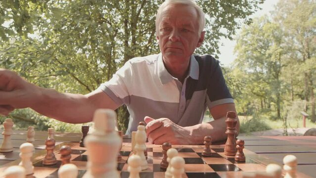 Waist up POV of concentrated Caucasian old man playing chess with male friend outdoors in park