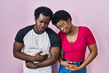Young african american couple standing over pink background with hand on stomach because indigestion, painful illness feeling unwell. ache concept.