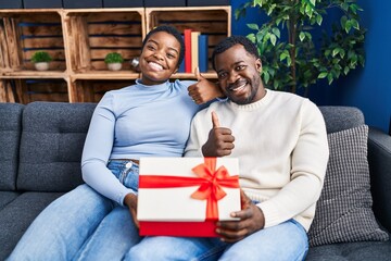 Young african american couple holding present smiling happy and positive, thumb up doing excellent and approval sign