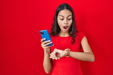 Young brazilian woman using smartphone over red background looking at the watch time worried,...