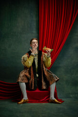 Yummy. Young man in vintage clothing eating hot dog against dark green, vintage background. Male...