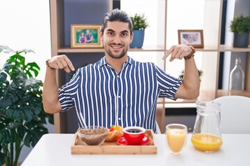 Hispanic man with long hair sitting on the table having breakfast looking confident with smile on face, pointing oneself with fingers proud and happy.