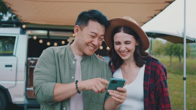 Multi-ethnic pair watching something hilarious on smartphone and laughing. Asian man and caucasian hipster girl enjoying funny video. Romantic summer road trip, outdoor activities