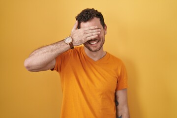 Young hispanic man standing over yellow background smiling and laughing with hand on face covering eyes for surprise. blind concept.