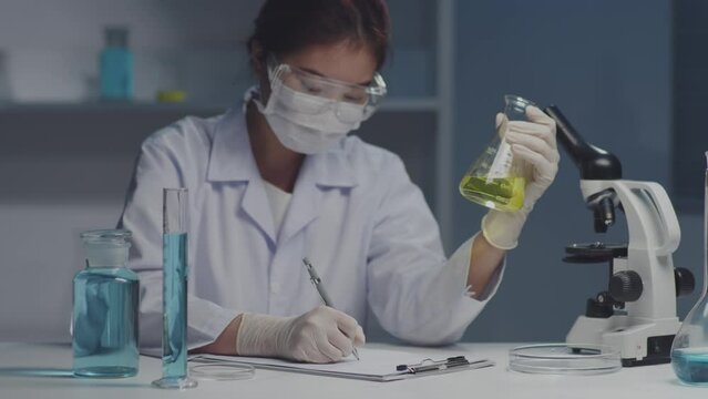 Scientist wearing white gloves and safety glasses is writing analysis of a chemical sample in the scientific research laboratory. Blue liquid contained inside some glassware on the table
