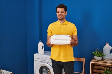 Young hispanic man holding clean towels at laundry room clueless and confused expression. doubt...