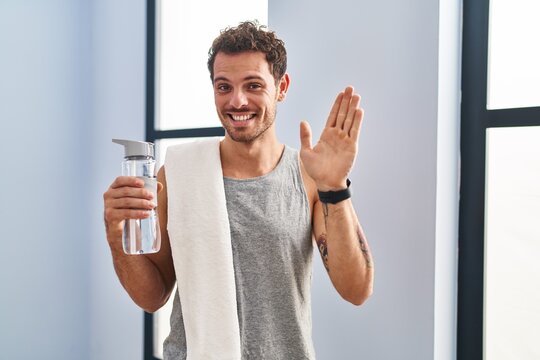 Young hispanic man wearing sportswear drinking water waiving saying hello happy and smiling, friendly welcome gesture
