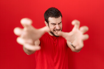 Young hispanic man wearing casual red t shirt shouting frustrated with rage, hands trying to strangle, yelling mad