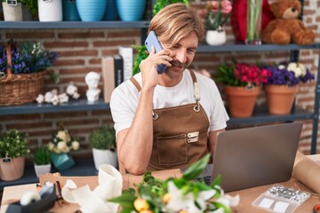 Young blond man florist talking on smartphone using laptop at flower shop