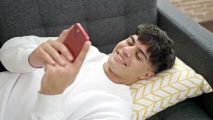 Obraz premium Young hispanic man using smartphone lying on sofa at home