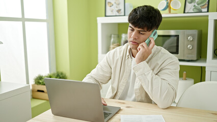 Naklejka premium Young hispanic man using laptop talking on smartphone at dinning room