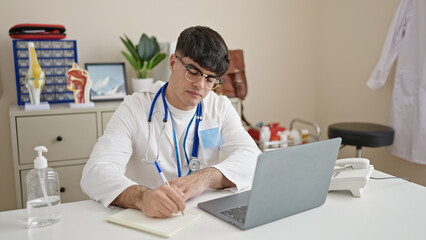 Young hispanic man doctor using laptop writing notes at clinic