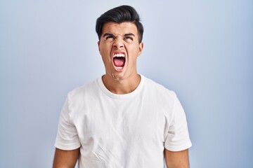 Hispanic man standing over blue background angry and mad screaming frustrated and furious, shouting with anger. rage and aggressive concept.