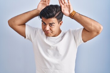 Hispanic man standing over blue background doing bunny ears gesture with hands palms looking cynical and skeptical. easter rabbit concept.