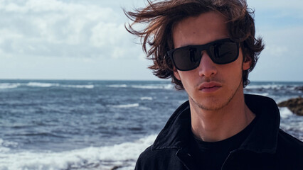 Young man with blown brown hair against the ocean on summer sunny day. Attractive young man looks...