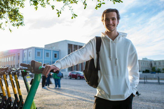 Student smiling while holding onto an electric scooter. Happy man standing close to an environmentally friendly vehicle. A cheerful young man with a backpack outdoors about to take a ride on a scooter