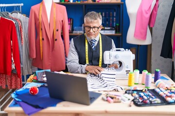 Middle age grey-haired man tailor using sewing machine and laptop at clothing factory