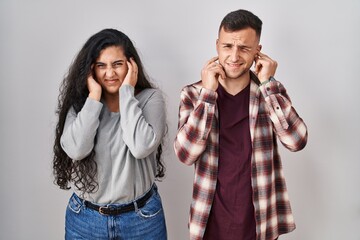 Young hispanic couple standing over white background covering ears with fingers with annoyed expression for the noise of loud music. deaf concept.
