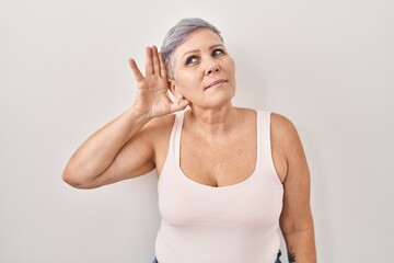 Middle age caucasian woman standing over white background smiling with hand over ear listening an hearing to rumor or gossip. deafness concept.