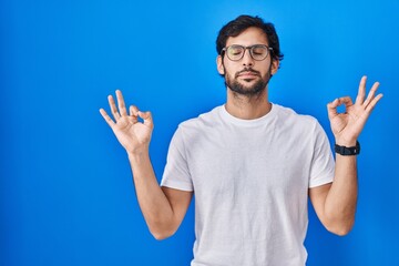 Naklejka premium Handsome latin man standing over blue background relax and smiling with eyes closed doing meditation gesture with fingers. yoga concept.