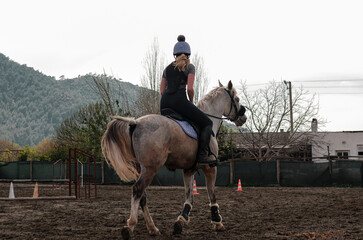 young european girl training horse, riding canter wearing back protection, gray gelding workout in...