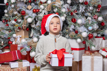 Cute happy child in santa hat holding a surprise gift on the background of a decorated Christmas...