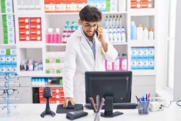 Young arab man pharmacist using computer talking on smartphone at pharmacy
