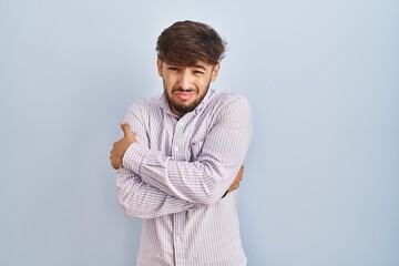 Arab man with beard standing over blue background shaking and freezing for winter cold with sad and...