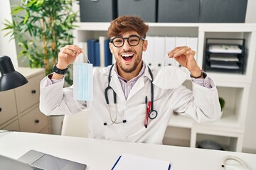 Arab man with beard wearing doctor uniform holding medical mask smiling and laughing hard out loud because funny crazy joke.