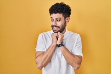 Arab man standing over yellow background laughing nervous and excited with hands on chin looking to...