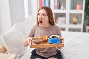 Obraz na płótnie Canvas Redhead woman wearing pajama holding breakfast tray angry and mad screaming frustrated and furious, shouting with anger. rage and aggressive concept.