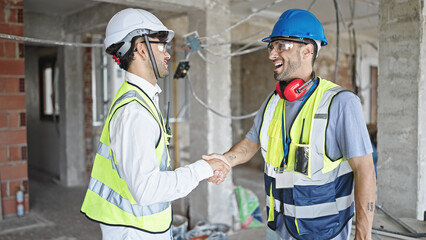 Two men builders smiling confident shake hands at construction site