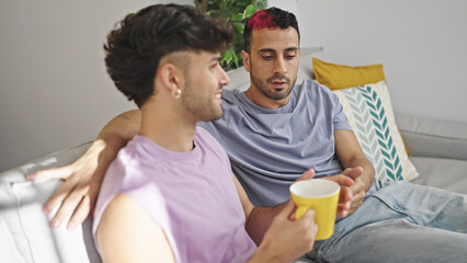 Two men couple drinking coffee sitting on sofa speaking at home