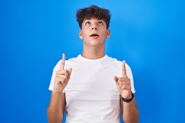 Hispanic teenager standing over blue background amazed and surprised looking up and pointing with fingers and raised arms.