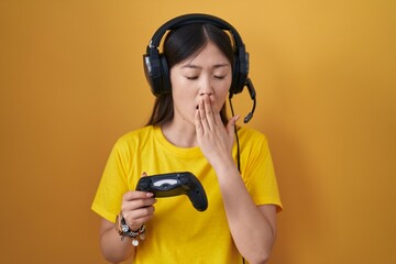 Chinese young woman playing video game holding controller bored yawning tired covering mouth with...