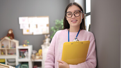 Young chinese woman preschool teacher smiling confident holding clipboard at kindergarten